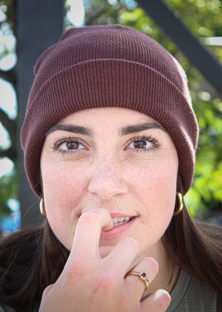 Emily Andrews, 21, wears a brown beanie and gold jewelry. She makes direct eye contact with the camera, posing hesitantly while biting her pointer finger.