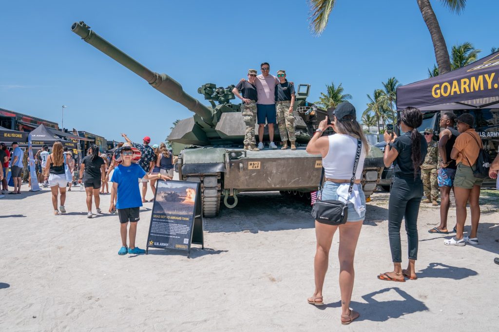 Corporal Simms and Specialist Valentine of the Third Infantry Division, 1st Armored Brigade Combat Team, 5th Squadron, 7th Cavalry Regiment pose with air show attendees on top of a M1A2 SEP-V3 Abrams tank in the display village of the Hyundai Air and Sea Show at Miami Beach on May 28, 2023.